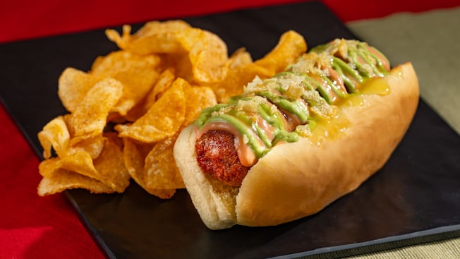 A large hot dog drizzled with condiments in a bun, sitting next to a serving of crisp potato chips