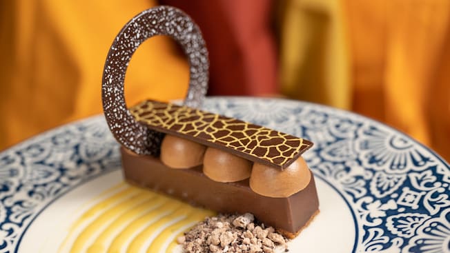 A plated dessert featuring a rectangular chocolate bar topped with round domes and a patterned chocolate ring, served with sauce and crumbled garnish
