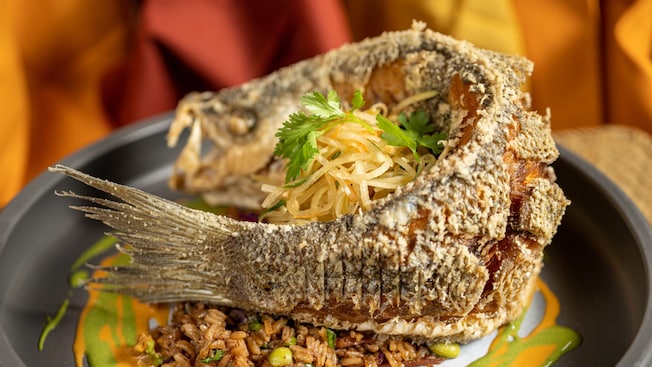 A whole fried fish presented with its head and tail intact, served atop a bed of rice and accompanied by a slaw and garnished with cilantro