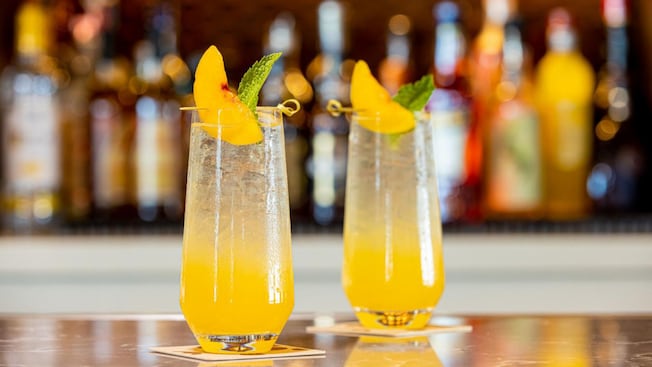 Two beverages garnished with fruit and mint sit on a bar