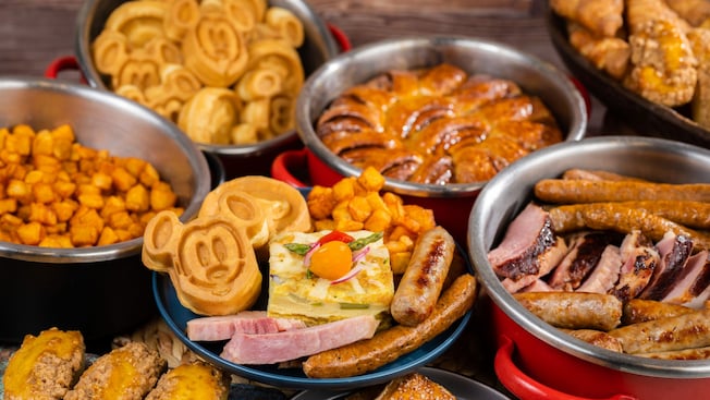 Un bufet de desayuno con waffles con forma de Mickey, salchichas, jamón, pastelitos y papas alargadas asadas