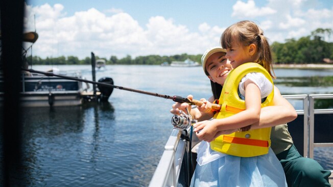 A girl holds a fishing pole in the water over the railing of a boat as her mother watches