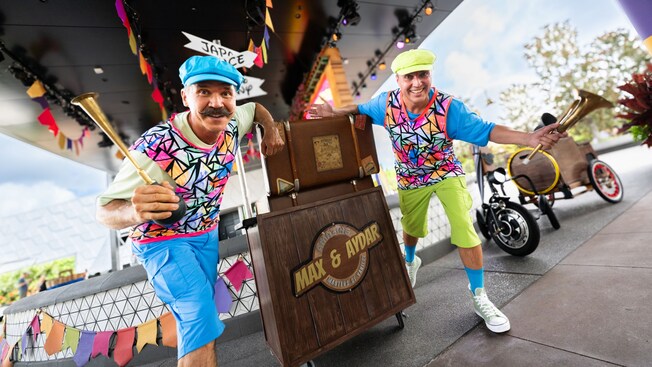 Los artistas de variedades Max y Aydar, con sus trompas en mano, posan junto a un podio y un triciclo de paseo en EPCOT	