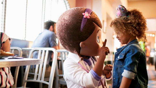 A young girl talking to Doc McStuffins in front of a dining table at Hollywood & Vine restaurant