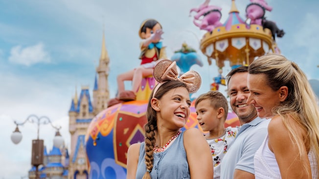 A smiling family in front of a parade float featuring Pinocchio and Dumbo, with Cinderella Castle in the background
