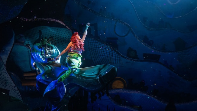 The Little Mermaid sings while perched on the edge of a large rock on the sea floor 