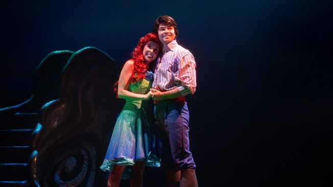 Princess Ariel and Prince Eric stand in front of a giant sea shell, holding hands