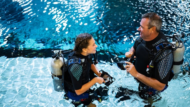 A teenage girl and her father, both in wetsuits and scuba gear standing waist deep in a pool   