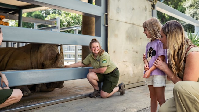 A zookeeper crouching beside a rhinoceros in a gated enclosure while a young girl and a woman look on