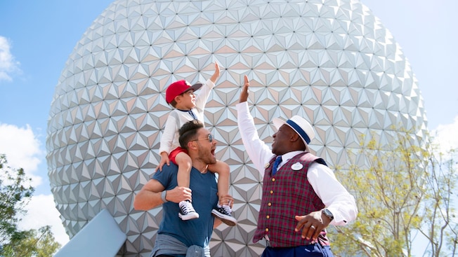 A young boy sitting on his father’s shoulders, giving a high five to a VIP Tour Guide in EPCOT