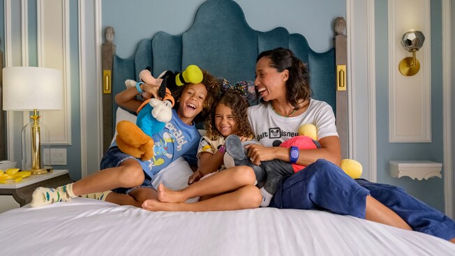 Una madre y dos niños sentados en una cama con peluches grandes de Goofy y Mickey Mouse en Disney's Grand Floridian Resort & Spa