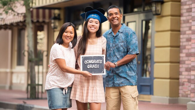 A young graduate, wearing a graduation cap that sports Mickey ears, is flanked on either side by her parents, who hold a sign in front of their daughter that reads, Congrats Grad