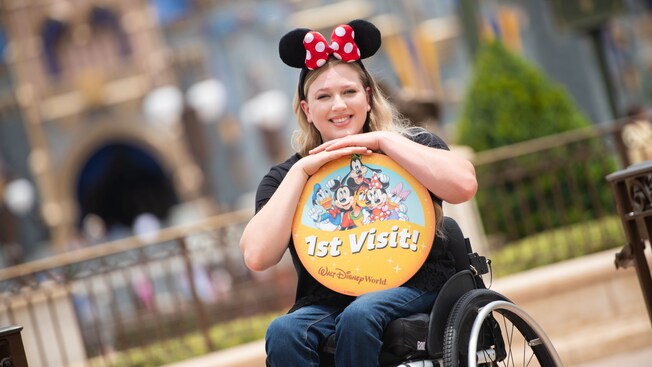 A smiling young woman, sitting in a wheelchair, wears a Minnie ears headband and holds a commemorative item featuring an image of the Fab Five and the words, First Visit, Walt Disney World