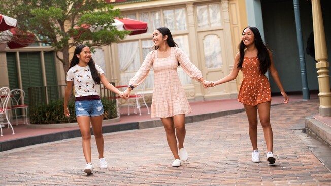 Three smiling young women hold hands as they walk, with outdoor tables and chairs in the background