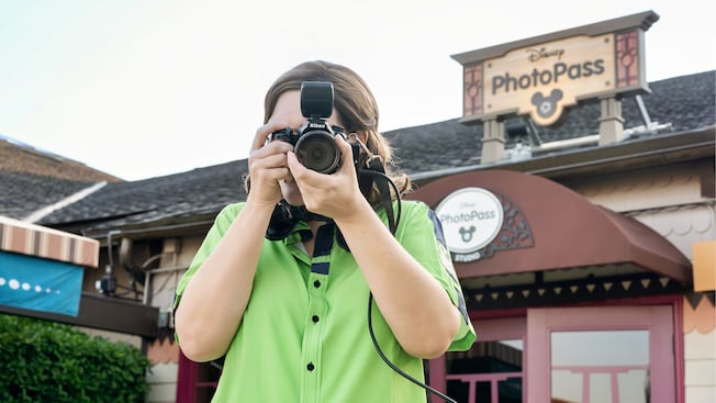 A woman stands outside a building with a sign that reads ‘Disney PhotoPass,’ taking a photo with her camera