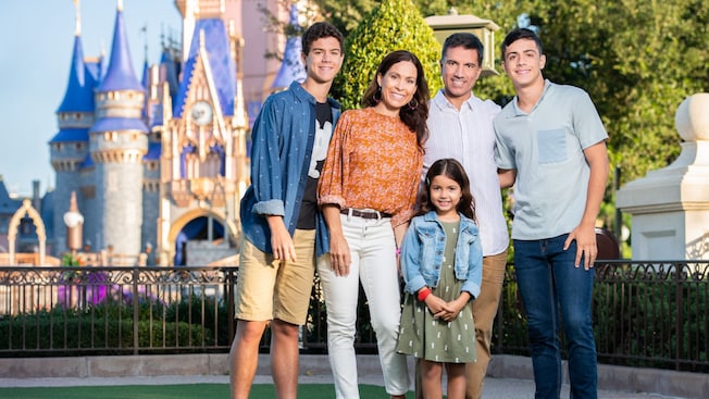 A family of 5 stands on a grass lawn in front of Cinderella Castle at Magic Kingdom park