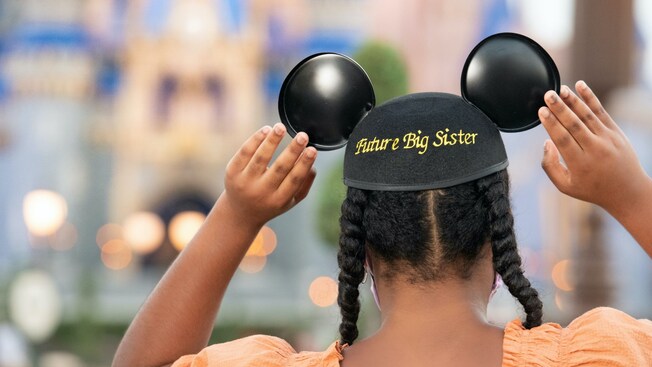 Seen from behind, a girl touches both ears of her Mickey ears hat, which is embroidered with the words, Future Big Sister