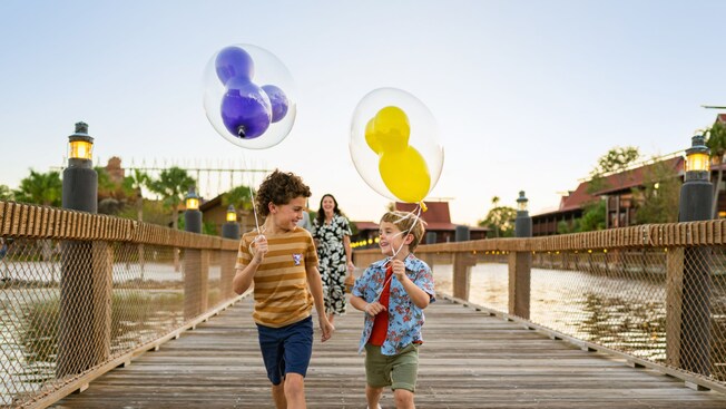Dos niños pequeños caminando por un malecón sosteniendo globos de Mickey Mouse