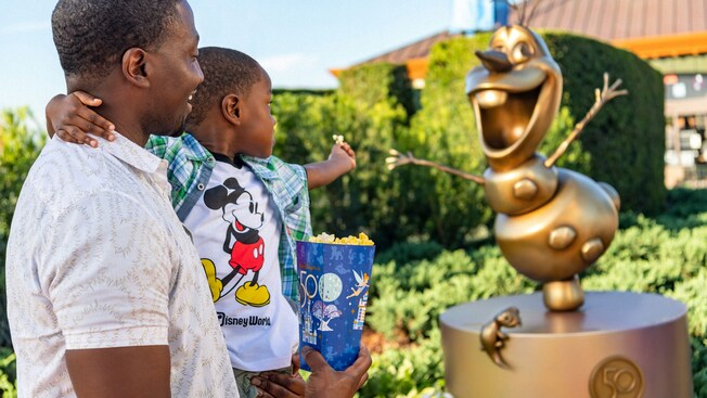 A man holds his son who holds a piece of popcorn out to a statue of Olaf