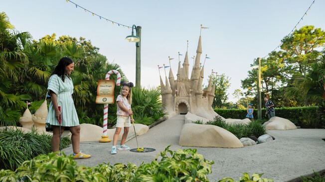 A mother and daughter playing miniature golf on a beach themed course at Winter Summerland Miniature Golf featuring a large sandcastle structure