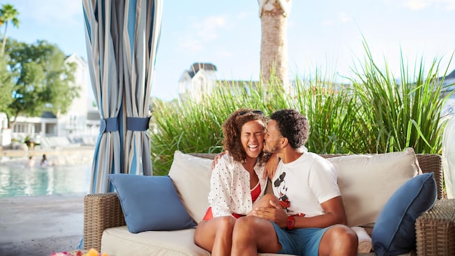 Un hombre besando a una mujer sonriente en la mejilla en un sofá de una cabaña junto a la piscina en un hotel Resort de Disney