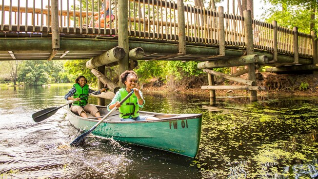 Una madre y su hija reman una canoa mientras pasan por debajo de un puente en Disney’s Fort Wilderness Resort and Campground