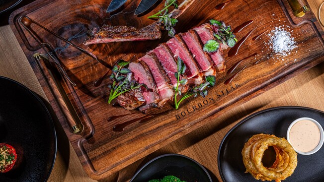 Una tabla de madera con un bistec Tomahawk cortado junto a una torre de onion rings y salsa para remojar en Bourbon Steak