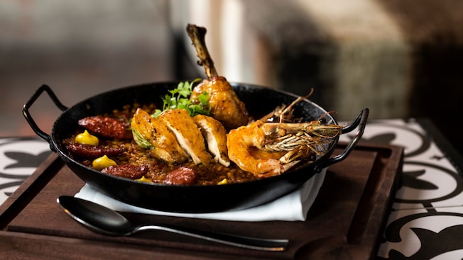 Una sartén de hierro fundido rellena de arroz con azafrán, pollo asado, camarones enteros y chorizo, servido sobre una tabla de madera