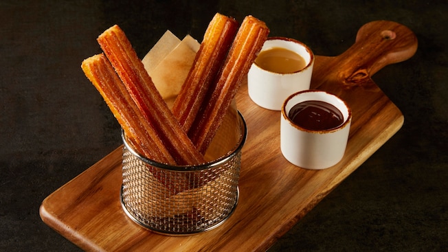 Churros con azúcar de canela en una cesta sobre una tabla con dos tazones de salsas