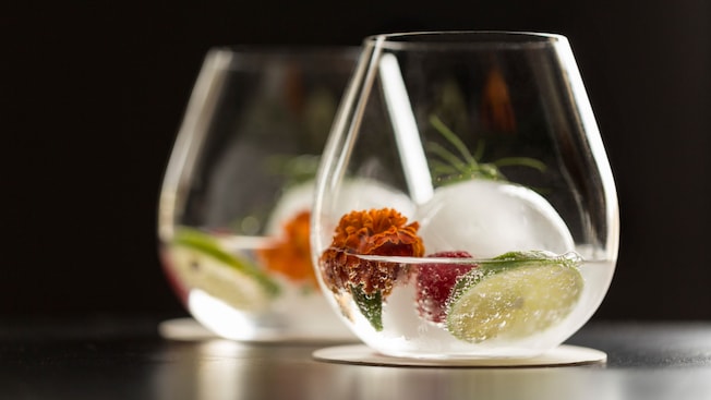 Dos copas sin tallo rellenas de cócteles y decoradas con limón verde, bayas, romero y hielo