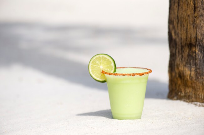 A cup of the Lime Dole Whip Margarita, sporting a lime slice on the cup’s rim, sits on a white sand beach