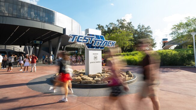 Guests walking past the Test Track entrance sign at Epcot as the monorail passes