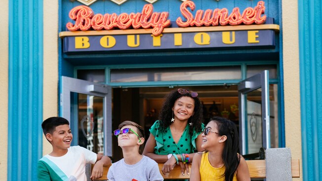 Four teens on a bench laughing in front of open doors and a sign with the words ‘Beverly Sunset Boutique’ 