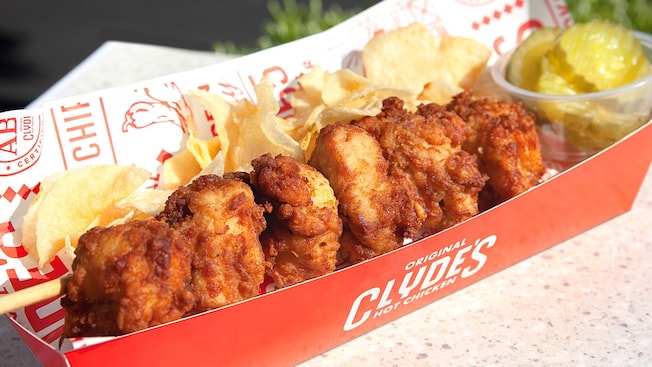 A stick with 5 pieces of breaded chicken on it, lying near pickle slices inside a paper hot dog tray