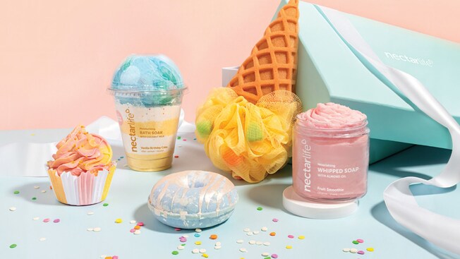 Una exhibición de productos de baño con temática de postre que incluye jabón en forma de pastelito, jabón batido en un jarro, un vaso con infusión de baño y una esponja de waffle con detalles de confeti