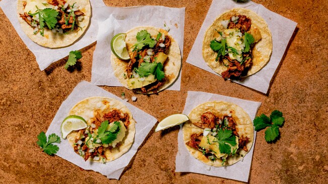 Ocho pequeños tacos al pastor adornados con limón verde y cilantro sobre en una mesa
