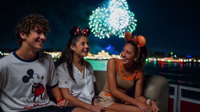 Three teens smiling on a fireworks cruise as fireworks burst above Seven Seas Lagoon