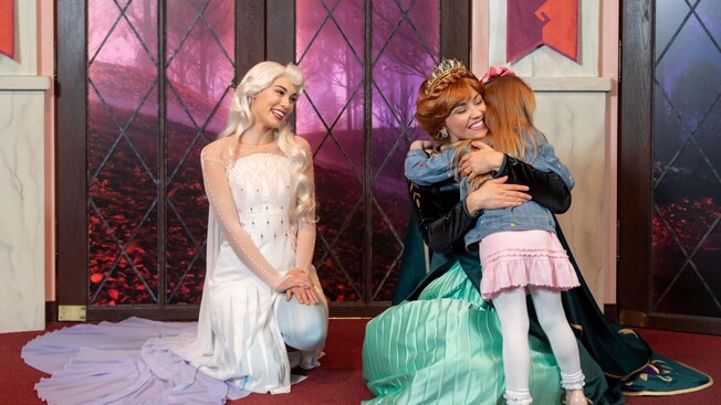 Anna crouches to hug a child as Elsa smiles at them