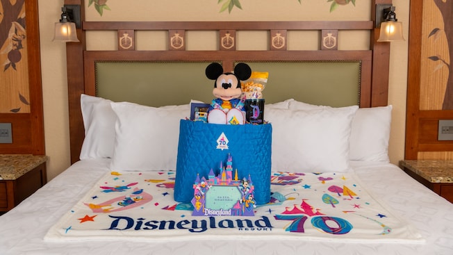 A Disneyland Resort 70th Celebration welcome basket, plush Mickey Mouse, photo frame and blanket on a Disneyland Resort hotel bed