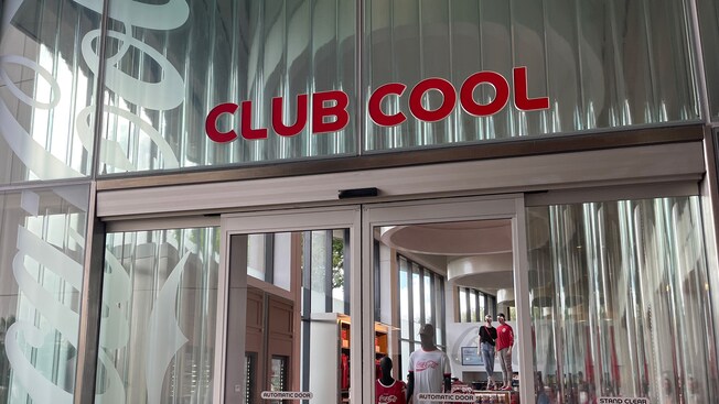 Un letrero de Club Cool sobre una puerta de vidrio y paredes de vidrio que muestran una gran sala con un letrero de Coca-Cola