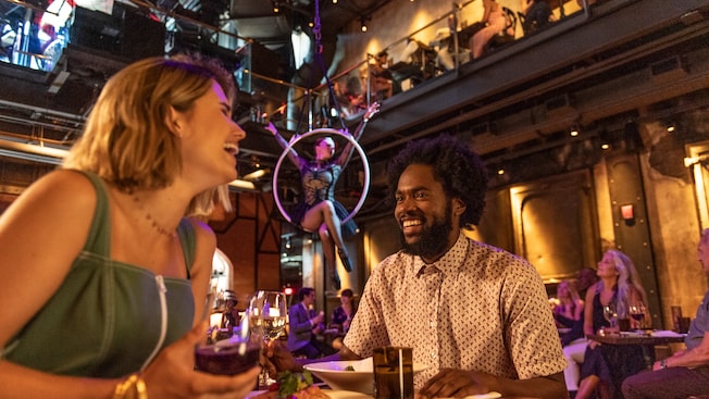 A couple at a dining table sharing a laugh while an aerialist balances on a hoop