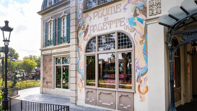 The exterior of Plume Et Palette in Epcot featuring a decorative mural with female figures reading, painting and the stylized name of the store on top