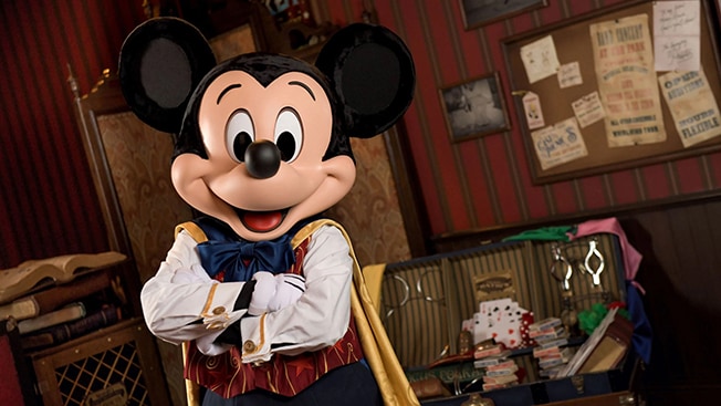 Mickey Mouse wears a cape and stands with his arms crossed in front of a large trunk and a pile of books 