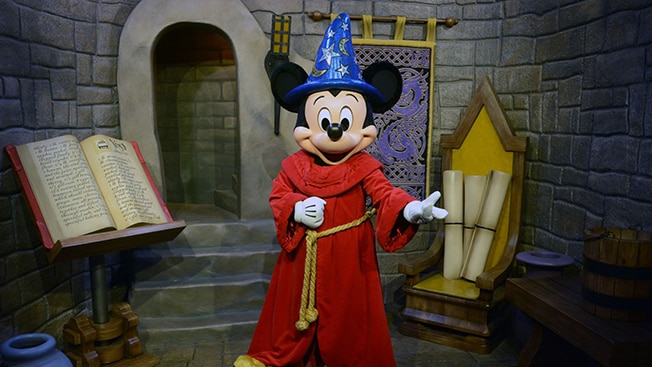 Mickey Mouse dressed as the Sorcerer’s Apprentice standing beside a large book inside a castle
