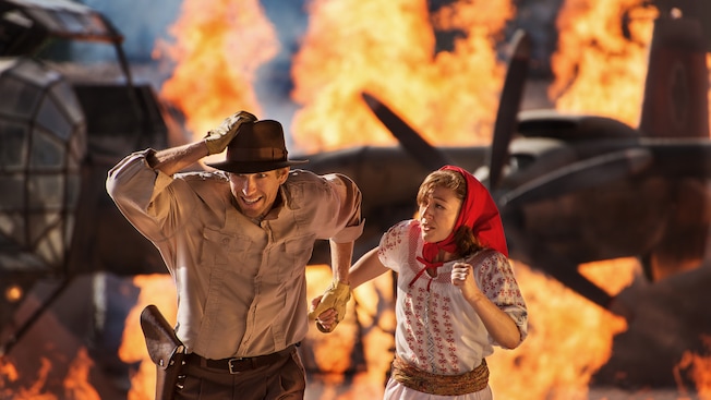 Performers as Indiana Jones and Marion Ravenwood run away from flaming airplanes