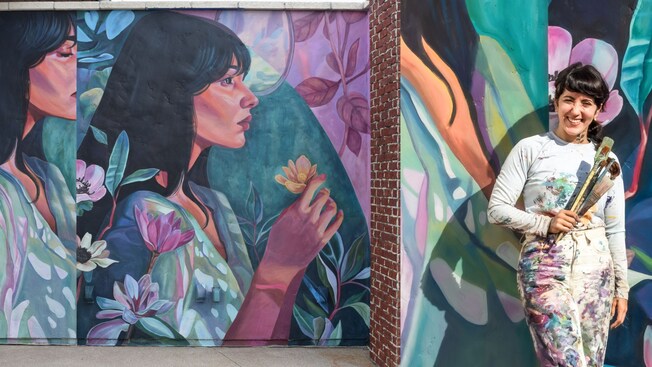Artist Nicole Salgar holding paintbrushes while standing in front of her mural portraying a woman with flowers
