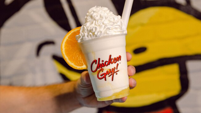 A person holding a Sunshine Shake topped with whipped cream and an orange slice from Chicken Guy