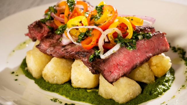 Steak topped with peppers, onions and herbs resting on a bed of potatoes and sauce