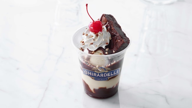 A plastic Ghirardelli cup holds ice cream, hot fudge, whipped cream, nuts, a brownie half and cherry