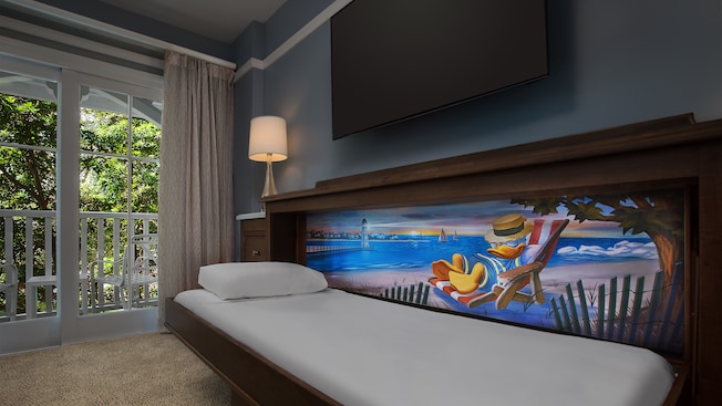A guestroom with a patio, lamp, TV and a pull down bed with a mural of Donald Duck lounging near the beach 